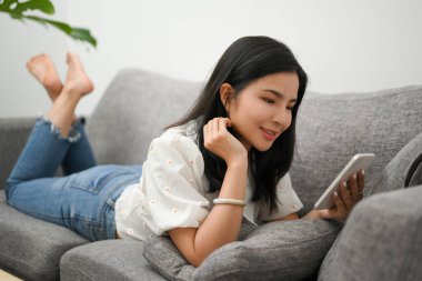 Çekici Asyalı genç kadın oturma odasında rahat bir koltukta uzanıyor, akıllı telefonuyla arkadaşlarıyla sohbet etmekten zevk alıyor..