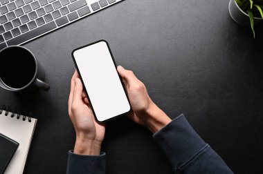 Üstten bakıldığında, koyu renk çalışma masasında cep telefonu kullanan bir erkek eli. Üst görünüm, telefon beyaz ekran modeli.