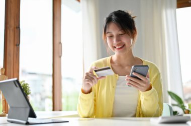 Çekici ve güzel Asyalı bin yıllık kadın masada bir kredi kartı ve akıllı telefon tutuyor. İnternetten alışveriş, internet pişirme, para transferi..