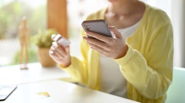 Asgari sarı hırkalı bir kadın ofis masasında, telefonda, parasını transfer etmek için internet bankacılığını kullanıyor. kırpılmış resim