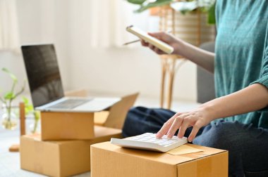 Görüntüyü kapat ve odakla, kadın çevrimiçi satıcı yerde oturuyor, teslimat paketlerini hazırlıyor ve hesap makinesi kullanıyor..