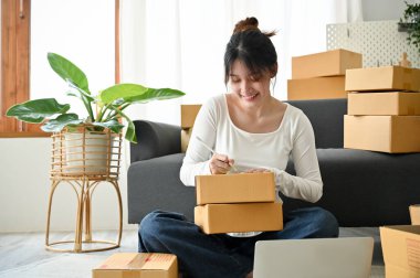 Startup small female business owner preparing her parcel boxes, writing the customer name and address on the box in her living room.