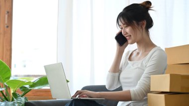 Successful asian female business startup entrepreneur talking on the phone with her supplier while using laptop computer.