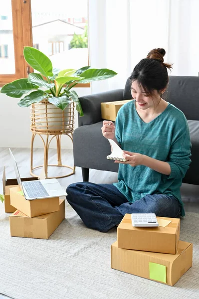 Portrait, Beautiful Asian millennial woman business startup entrepreneur notes something on a spiral notepad while preparing the delivery boxes.