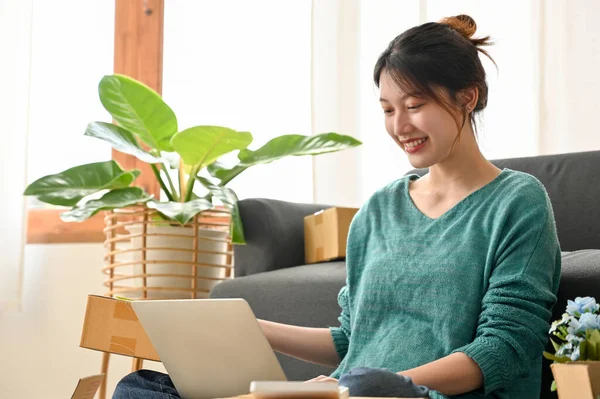 Pretty Asian millennial woman using laptop computer to update her product packing status to her customers in living room.
