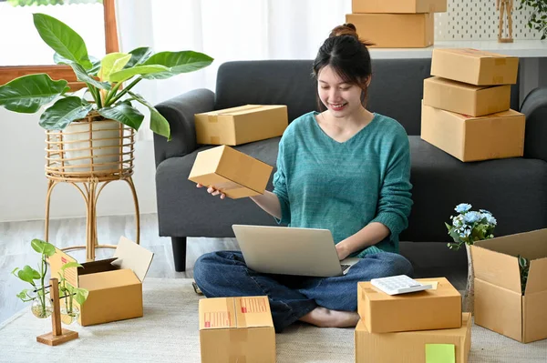 Attractive Asian female retail business owner using laptop computer, updating her product delivery status and preparing the product to ship.