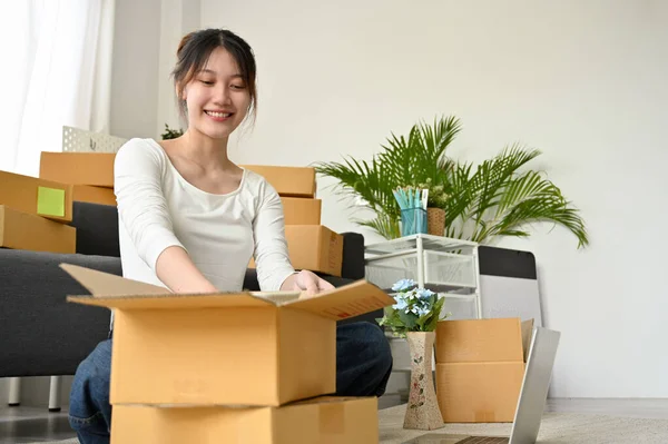 Attractive Asian young woman packing her stuff in a cardboard boxes, preparing parcel for relocate or moving into new home.
