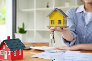 Female real estate agent holding or showing a house model and house keys over the office desk. Rental or property investment concept, closeup