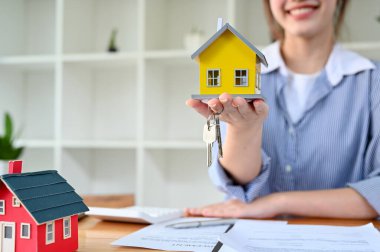 Beautiful Asian female house insurance broker or real estate agent realtor holding a yellow house model at her desk. cropped image