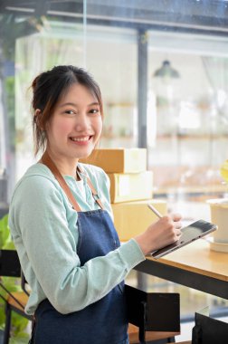 Güzel ve neşeli Asyalı genç bir kadının portresi çevrimiçi dükkan sahibi üniforması iş yerinde duruyor ve elinde bir tablet dokunmatik ped var.