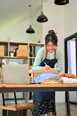 Neşeli Asyalı bin yıllık kadın giyim mağazası sahibi paket kutusu hazırlıyor, kıyafetlerini müşterilere gönderiyor. KOBİ iş kavramı.