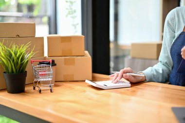 Kadın dükkan sahibi, atölyesindeki ürün maliyetini ve çevrimiçi pazarlama maliyetini kontrol etmek için hesap makinesi kullanıyor. kırpılmış, kapatılmış resim