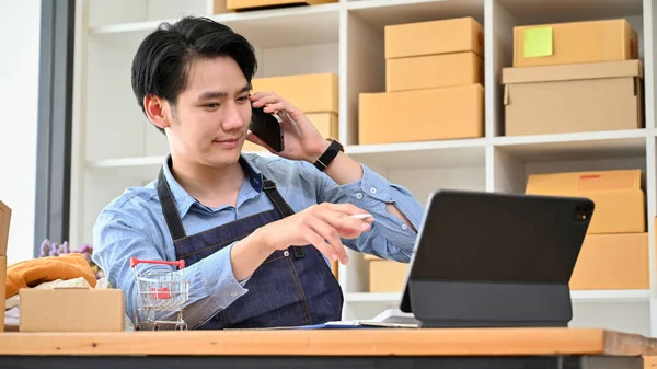 Handsome Asian millennial male small online business entrepreneur talking on the phone while looking some information on tablet screen.
