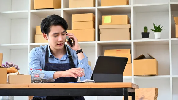 Smart Asian millennial male startup business owner talking on the phone with his material supplier while working at his desk.