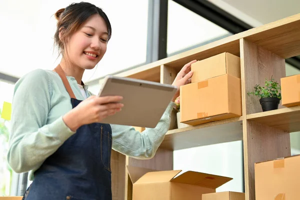 Kadın dükkan sahibi dijital tablet kullanıyor ve depolama odasındaki bir paket kutusunda ürün stoklarını kontrol ediyor..