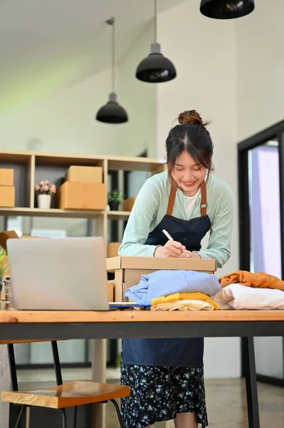 Neşeli Asyalı bin yıllık kadın giyim mağazası sahibi paket kutusu hazırlıyor, kıyafetlerini müşterilere gönderiyor. KOBİ iş kavramı.