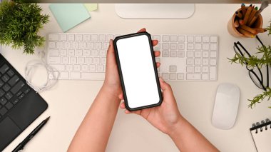 Smartphone white screen mockup on a female's hands over modern white office desk workspace. top view