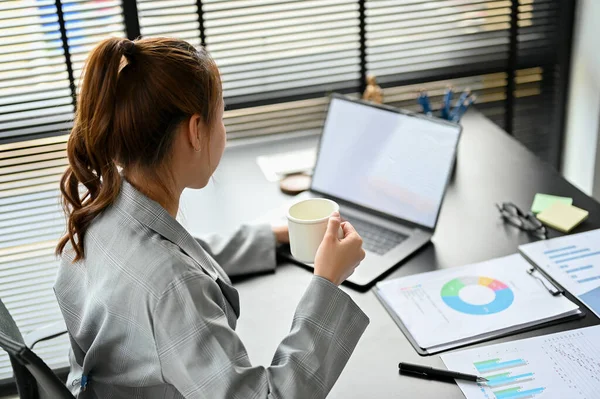 Successful Asian millennial businesswoman or entrepreneur sipping a morning coffee while working on laptop computer at her desk.