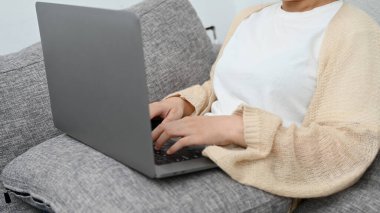 A charming Asian young female relaxes on sofa in her living room and using laptop computer. Remote working at home