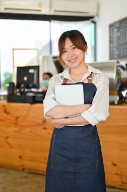 Portrait of an Attractive Asian young female barista standing in front of the counter and holding a tablet. Coffee shop worker, Entrepreneur, Barista.