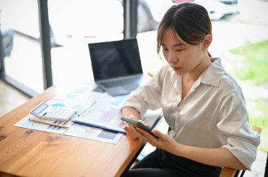 Bin yıllık güzel ofis çalışanı masada oturuyor ve ofisteki cep telefonunu kullanıyor..
