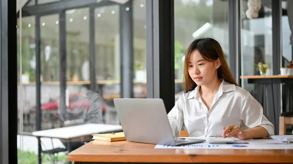 Concentrated Asian female financial analysts or businesswoman reviewing a financial data report and using laptop computer in the office.