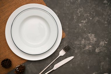 Lüks yemek masası seti kahverengi servis altlığında beyaz seramik tabak, şık koyu arka planda gümüş çatal bıçak takımı. en üstteki görünüm