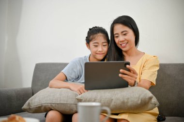 Mutlu Asyalı anne ve küçük kızı oturma odasında rahat bir kanepede oturup tablet üzerinde çocuk çizgi filmleri izliyorlar..