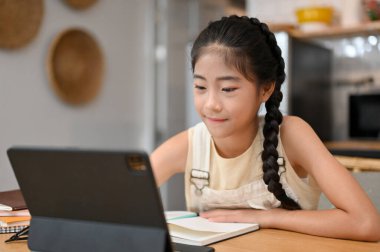 Güzel Asyalı küçük kız yemek odasında oturmuş online ders çalışıyor ve salgın sırasında online ders alıyor. Çocuklar için ev okulu kavramı.