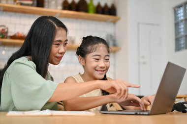 Neşeli Asyalı anne dizüstü bilgisayara bakıyor, kızına online ödevinde yardım ediyor, soruyu açıklıyor ve cevabı söylüyor..