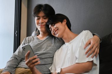 Eşcinsel bir adam, erkek arkadaşının omzuna sarılıp oturma odasında rahatlıyor ve birlikte akıllı telefondan bir şeyler seyrediyor. LGBT çiftler konsepti