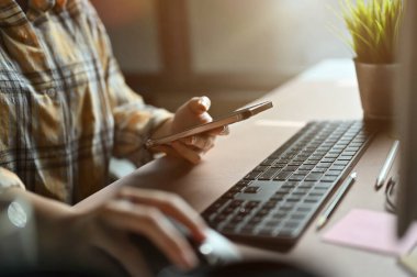 Yakın çekim, ofis masasında çalışan kadın ofis çalışanı, elinde akıllı telefon ve bilgisayar kullanıyor..