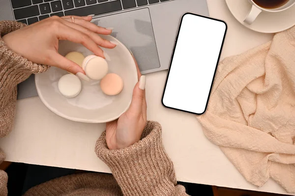 Top view, Female officer eating a macaron while working at her office desk. Workspace with laptop and smartphone blank screen mockup.