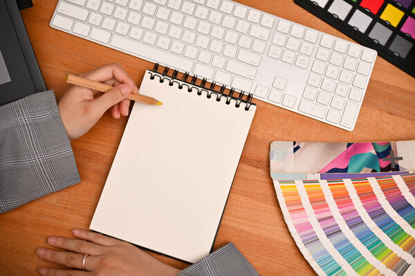 Graphic designer office desk with a female hand writing something on blank spiral notepad. top view image