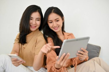 İki güzel Asyalı kadının portresi oturma odasında dinleniyor, dijital tablet ve akıllı telefon kullanıyor, birlikte bazı çevrimiçi video içeriklerine bakıyorlar..