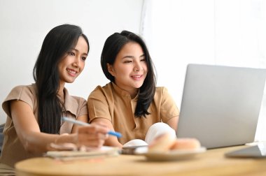 Modern, parlak kafede birlikte oturan iki çekici Asyalı genç kadın dizüstü bilgisayar kullanarak ekrana bakıyorlar. Uzak çalışma konsepti