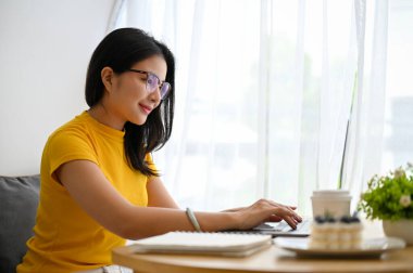 Gözlüklü çekici Asyalı kadın modern, parlak çalışma odasında oturuyor, projesini kuruyor, dizüstü bilgisayar kullanıyor..