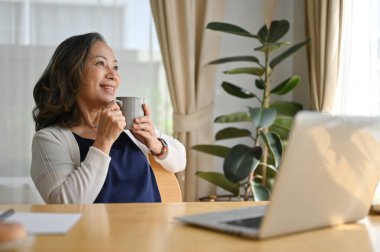 Gülümseyen, orta yaşlı, şık bir kadın. Sabahları modern oturma odasındaki koltukta dinlenmekten zevk alıyor..