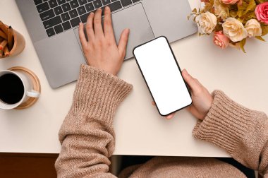 Modern akıllı telefon kullanan ve kadınsı çalışma masasında dizüstü bilgisayarda klavye kullanan bir kadının üst görüntüsü. Telefon boş ekran modeli