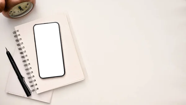 Minimal white office desk with smartphone white screen mockup on spiral notepad, pen, vintage clock and copy space on white background. top view