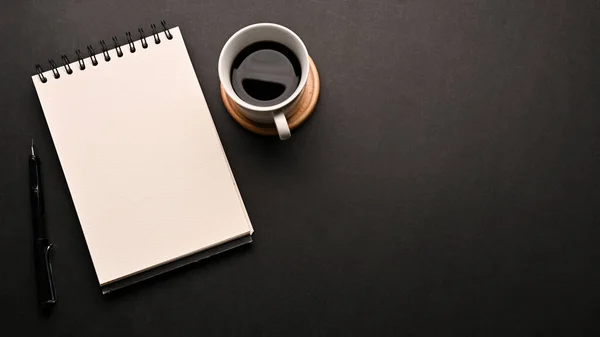 Minimal black workspace table with a cup of coffee, an empty spiral notepad and copy space on black background. above view