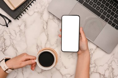 Görüş alanının üstünde, kadın akıllı telefon kullanırken modern beyaz mermer çalışma masası arka planında bir sabah kahvesini yudumluyor. Telefon boş ekran modeli.