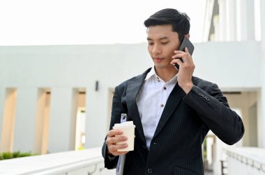 Zeki ve yakışıklı Asyalı iş adamı ya da resmi takım elbiseli erkek bankacı, bina boyunca yürüyor, telefonla konuşuyor ve elinde kahve fincanı tutuyor..