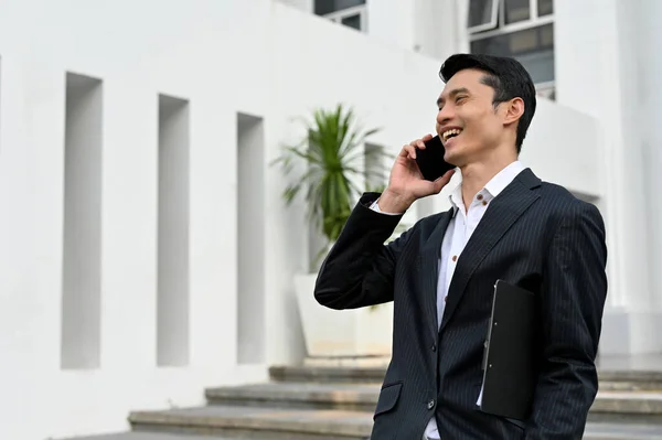 Cheerful asian businessman in formal suit, standing outside of the building and talking on the phone with business client