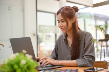 Yaratıcı genç Asyalı grafik tasarımcı ofis masasında oturuyor, görevi üzerinde çalışıyor ve taşınabilir dizüstü bilgisayar kullanıyor..