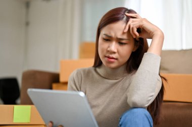 Stresli Asyalı genç bayan alışveriş mağazası sahibi tabletten siparişlerini kontrol ediyor ve müşterilerin yorumlarını kontrol ediyor..