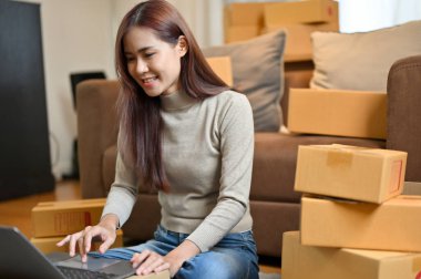 Başarılı ve neşeli Asyalı genç bayan alışveriş mağazası sahibi online siparişlerini kontrol ediyor ve dizüstü bilgisayardaki ürün durumunu güncelliyor..