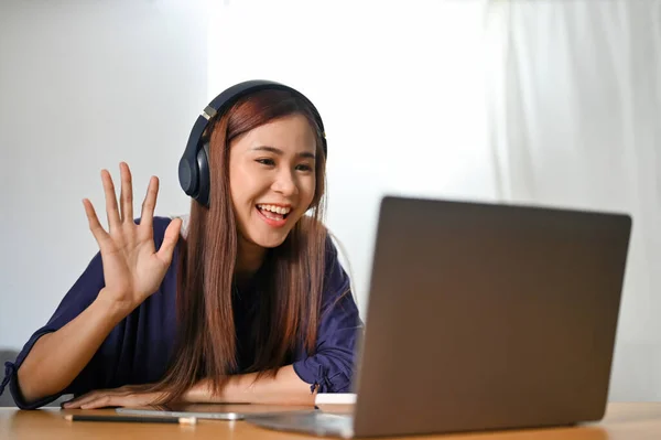 Cheerful and attractive Asian female tutor is tutoring her students on online meeting via laptop computer. A friendly young Asian woman is talking with her foreign friends on webcam. Hi, Hello gesture