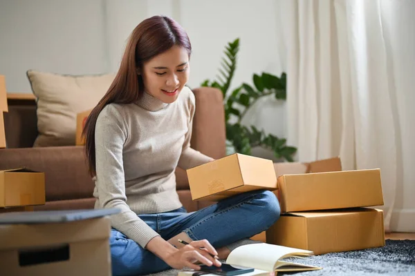 Cheerful young asian female online shop owner in the living room preparing her parcel boxes, be ready to ship to her customers.