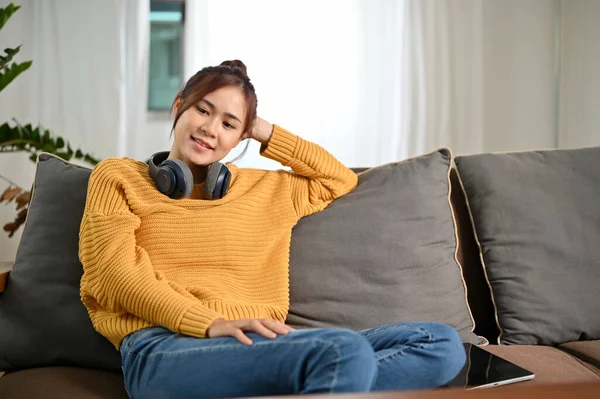 リビングルームで快適なソファに座っている間 現代的なワイヤレスヘッドフォンリラックスを持つ美しく魅力的な若いアジアの女性 — ストック写真